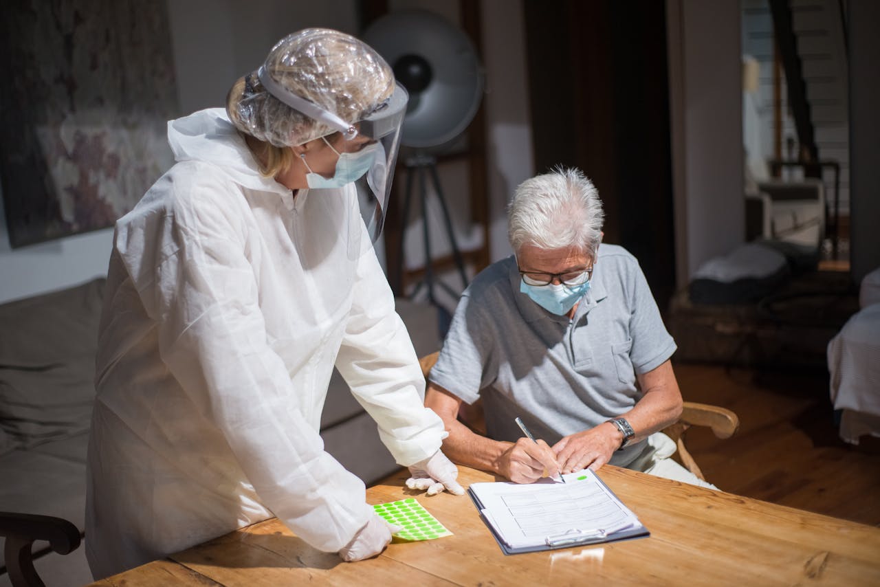 A healthcare worker in protective gear assists an elderly man with paperwork in a home setting.